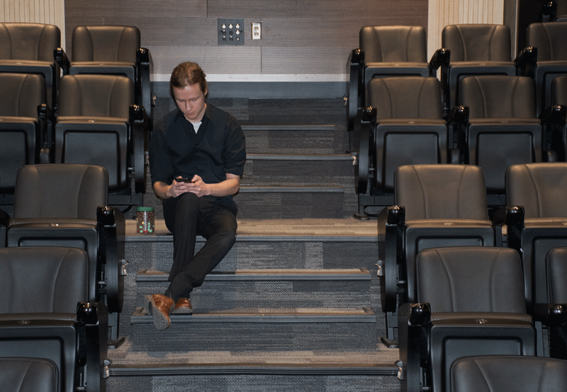 Photograph of a young man using a cellphone while sitting on the stairs surrounded by theatre seats