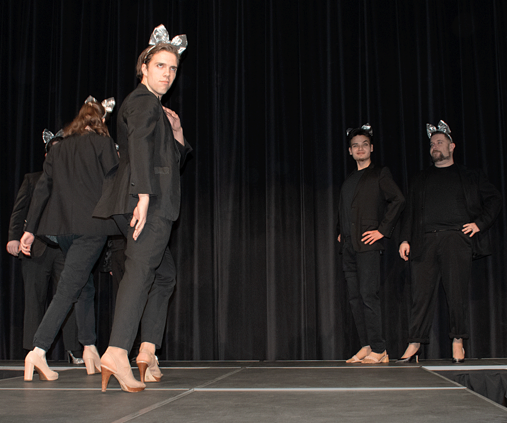 A group of male models posing in high heels and wearing sparkly silver bows in their hair.
