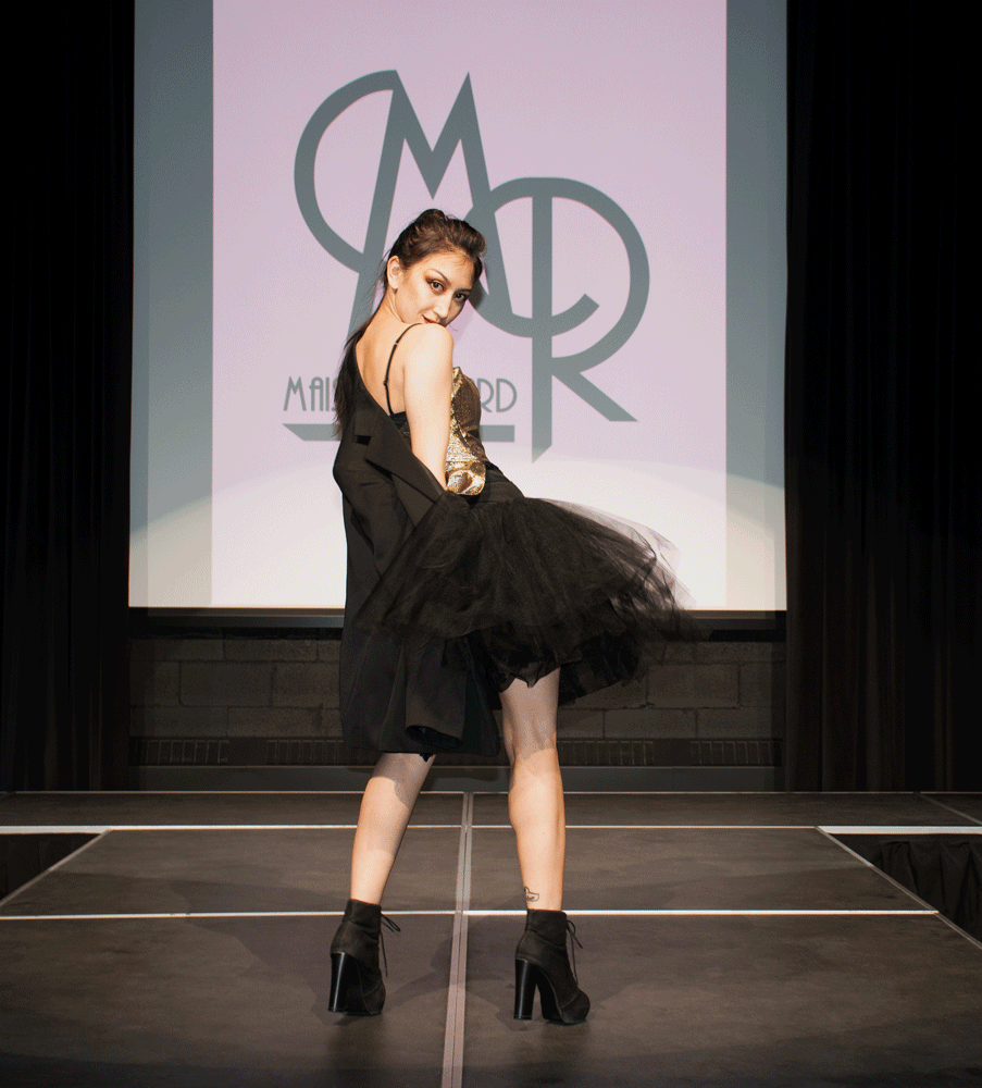 A young woman in a black dress on a runway, her back to the camera and giving a sassy look over her shoulder.