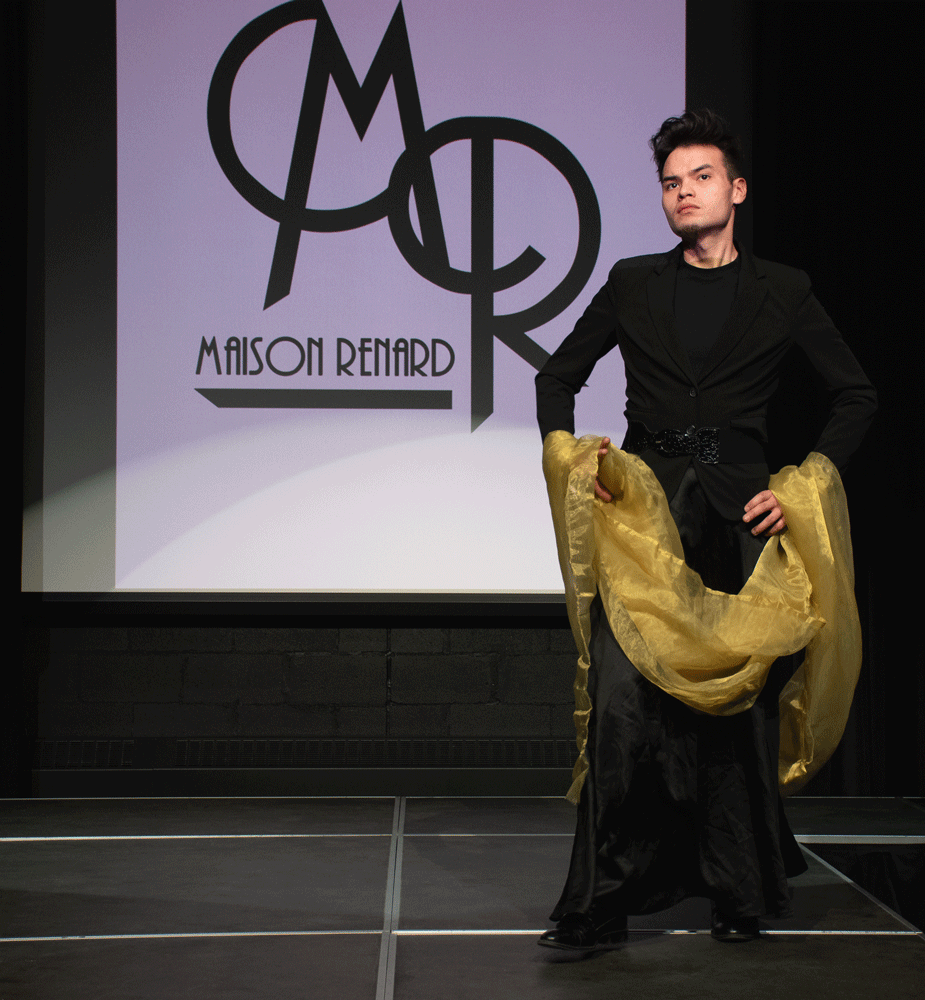 A tall man wearing a black dress with a scarf of gold fabric over his arms.