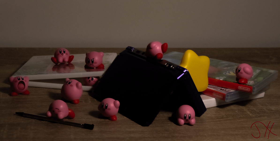 Several figurines of video game character Kirby posed around a Nintendo 3DS. Kirby's super star is crashed into the back of the Nintendo 3DS.