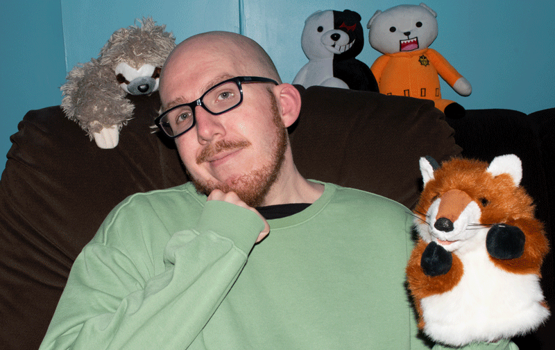 A photo of Jarin Helliwell sitting in an easy chair with a fox puppet on one hand, a sloth plushie over his shoulder, and plushies of Bepo from One Piece and Monokuma from Danganronpa on the couch in the background